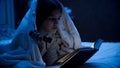 Portrait of cute girl covering with blanket and reading book with light torch