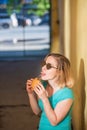 Portrait of a cute girl in a green dress walking outside and eating a burger on a warm summer day. Beautiful blonde in sunglasses