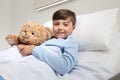 Portrait of cute happy child alone in hospital room lying in bed with teddy bear looking at camera and smiling with copy space