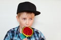 A portrait of a cute little blond boy in black cap and checked shirt eating a bright lollipop isolated over white background looki Royalty Free Stock Photo