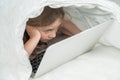 Portrait of a cute little girl using a laptop while lying under a blanket. Royalty Free Stock Photo