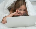 Portrait of a cute little girl using a laptop while lying under a blanket. Royalty Free Stock Photo