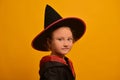 Little wizard girl in Halloween costume on yellow background. Kid in magic hat and mantle or robe looking at camera.