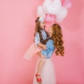 Portrait of cute long-haired birthday girl with white helium balloons embracing her young curly mom after event. Mother