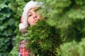 Portrait of cute lovely gir in a santa claus red hat sending blowing kiss with pout lips looking at camera through green branches. Royalty Free Stock Photo