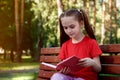 Portrait of cute pensive little girl sitting on the wooden bench with open book in her hands outdoors Royalty Free Stock Photo