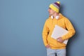 Portrait of cute positive man wearing beanie hat and casual yellow hoodie standing isolated over blue background, looking away at Royalty Free Stock Photo