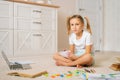 Portrait of cute primary girl child writing homework in notebook sitting on floor at home, looking at camera. Royalty Free Stock Photo