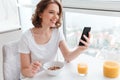 Portrait of cute smiling girl in white tshirt chatting on smartphone while sitting and eating cornflakes at the kitchen table Royalty Free Stock Photo