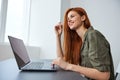 portrait of a cute, smiling red-haired woman working at a laptop while sitting at an empty table in a bright room Royalty Free Stock Photo