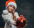 Teen boy wearing Santa`s hat holding a big Christmas ball in a dark room with a decorated wooden ladder. Royalty Free Stock Photo