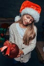 Teen boy wearing Santa`s hat holding a big Christmas ball in a dark room with gifts. Royalty Free Stock Photo