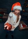 Teen boy wearing Santa`s hat holding a big Christmas ball in a dark room with gifts. Royalty Free Stock Photo