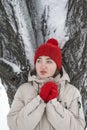 Portrait of cute thoughtfully girl looking to the side. Young woman in red hat and mittens stands near snow-covered tree