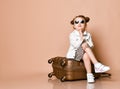 Stylish baby kid girl in leopard print dress, denim jacket and sunglasses is sitting on roller suitcase at copy space Royalty Free Stock Photo