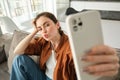 Portrait of cute young woman, modern girl taking selfies on smartphone app, posing on couch, extends her white mobile Royalty Free Stock Photo