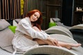 Portrait of cute young woman wearing white bathrobe relaxing sitting on deck chair by poolside after beauty treatment at Royalty Free Stock Photo