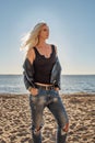 A portrait of a daring blonde girl in a tight-fitting t-shirt and torn jeans standing on the sandy beach of the sea and looking th Royalty Free Stock Photo