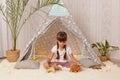 Portrait of dark haired cute little girl playing with toys and in teepee tent, sitting on floor on soft carpet, posing alone in Royalty Free Stock Photo
