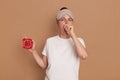 Portrait of dark haired young adult woman in cat sleeping eye mask yawning and covering mouth with hand, holding red alarm clock Royalty Free Stock Photo