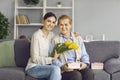 Happy daughter and senior mom sitting on sofa with presents and flowers on Mother& x27;s Day