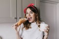 Portrait of debonair young woman eating croissant with surprised smile. Elegant curly girl posing i