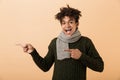 Portrait of delighted african american guy wearing sweater and scarf gesturing aside, isolated over beige background