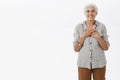 Portrait of delighted charming old female with grey combed hair holding palms on chest with pleased and grateful smile