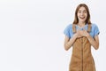 Portrait of delighted greatful charming kind young girl in brown overalls holding palms pressed to chest in thankful