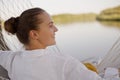 Portrait of delighted happy dark haired woman wearing white shirt sitting in hammock on the bank of the river or lake, looking Royalty Free Stock Photo