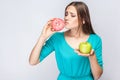 Portrait of desiring young beautiful girl in blue blouse standing, holding pink donut and green apple in the hands and wanted