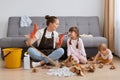 Portrait of despaired woman with bun hairstyle wearing white t shirt, brown apron and jeans, cleaning house, sitting on floor near