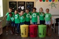 Group of schoolchildren holding color coded recycling bins and bags in an elementary school classro