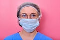 Portrait of a doctor woman in glasses and a medical hat, closeup. Nurse in a blue uniform and a protective mask on a pink