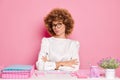 Portrait of doubtful business woman with curly Afro hair sits at white desktop with papers, looks thoughtful dreams