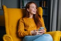 Portrait of dreaming redhead young woman using mobile phone sitting in yellow armchair, smiling looking away. Royalty Free Stock Photo