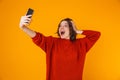 Portrait of ecstatic woman holding and taking selfie photo on cell phone while standing isolated over yellow background Royalty Free Stock Photo