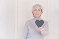 A portrait of an elderly Caucasian woman with short gray hair wearing glasses holding in her hand a black heart.