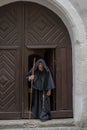 Portrait of an elderly hunched monk 45-50 years old with a beard in a black cassock with a staff coming out of the textured gate o Royalty Free Stock Photo