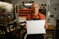 Portrait of elderly man with empty blank mock-up poster. Male mechanic in garage interior or workshop. Bike service Royalty Free Stock Photo