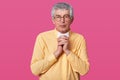 Portrait of elderly man with rounded spectacles, looks seriously at camera and keeps fingers crossed. Mature male dressed in Royalty Free Stock Photo