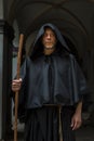 Portrait of an elderly monk 45-50 years old with a beard in a black robe with a staff, standing under the arches of a monastery or Royalty Free Stock Photo