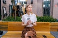 Portrait of elegant blonde young woman sitting on bench holding paper cup of takeaway coffee pensive looking away. Royalty Free Stock Photo