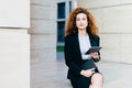 Portrait of elegant lady dressed in white blouse, black jacket and skirt, holding tablet and pocket book while sitting near office