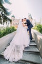 Portrait of elegant stylish young wedding couple kissing on stairs in park. Romantic antique palace at background