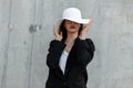 Portrait elegant young woman in glasses in summer beautiful hat who covers face in stylish black blazer near vintage wall on Royalty Free Stock Photo