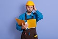 Portrait of emotional magnetic helpless young builder looking directly at camera, holding papercase, putting one hand on helmet,