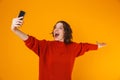 Portrait of emotional woman holding and taking selfie photo on cell phone while standing isolated over yellow background Royalty Free Stock Photo