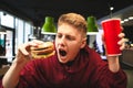 Portrait of an emotional young man holding a glass of beverage and a burger,looking at a burger and going to bite