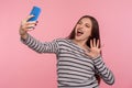 Portrait of enthusiastic woman in striped sweatshirt taking selfie on mobile phone and smiling crazy